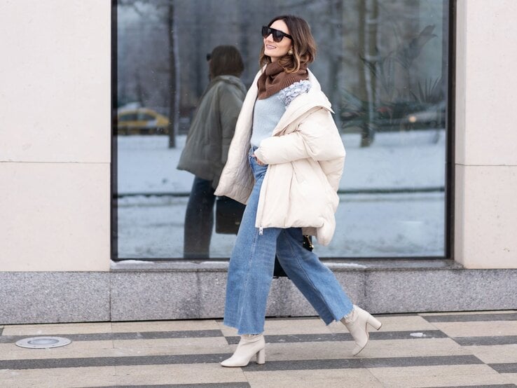 Modebewusste Frau in heller Steppjacke, Jeans und beigen Stiefeletten spaziert bei Winterwetter vor einer Glaswand entlang.