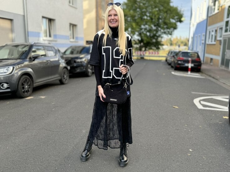 Frau in einem sportlich-modischen Outfit, das an den Blokecore-Trend angelehnt ist. Sie trägt ein übergroßes, schwarz-weißes Jersey mit der Nummer "13", kombiniert mit einem langen Spitzenrock und robusten, schwarzen Stiefeln.