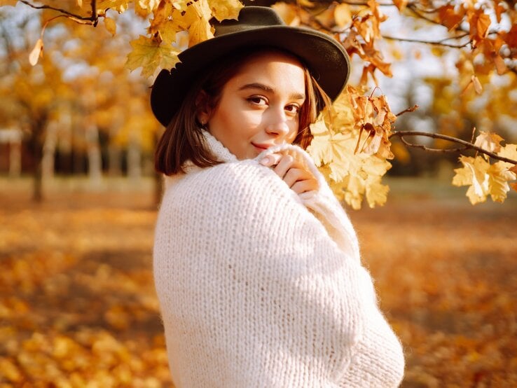 Junge Frau mit schwarzem Hut und weißem Strickpullover lächelt leicht inmitten herbstlicher gelber Blätter.