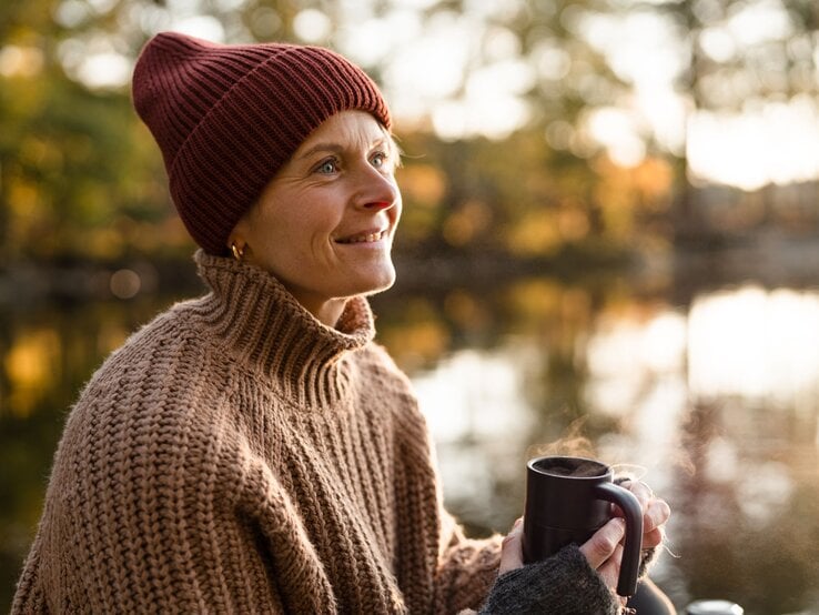 Lächelnde Frau in braunem Pullover und Mütze genießt warmes Getränk am Morgen, umgeben von herbstlicher Natur.