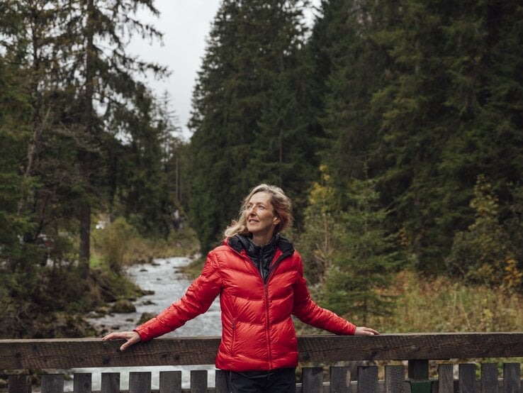 Frau mittleren Alters in roter Jacke lehnt lächelnd an einer Holzbrücke, umgeben von dichtem Wald und Bachlauf.