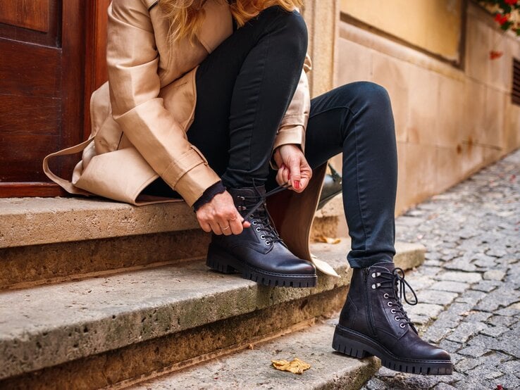 Nahaufnahme einer Frau in schwarzer Jeans und beigem Mantel, die ihre robusten Schnürstiefel schnürt.