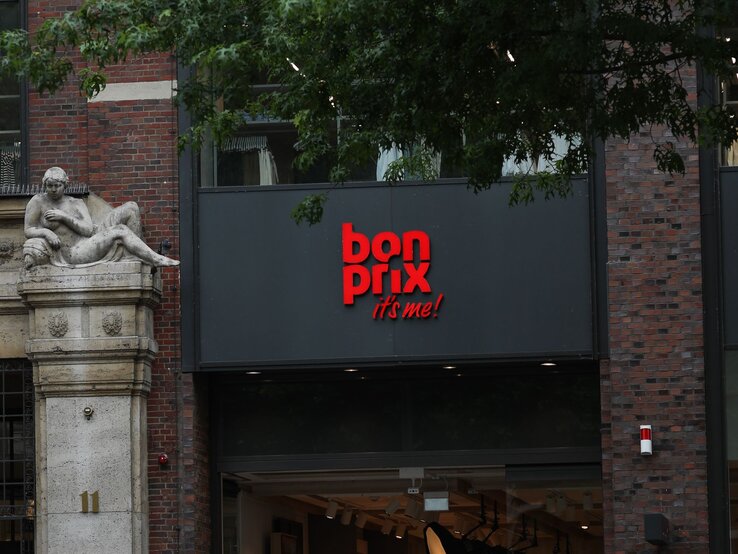 Bonprix-Logo in leuchtendem Rot auf einer grauen Fassade, daneben eine historische Steinskulptur vor Backsteinmauer.