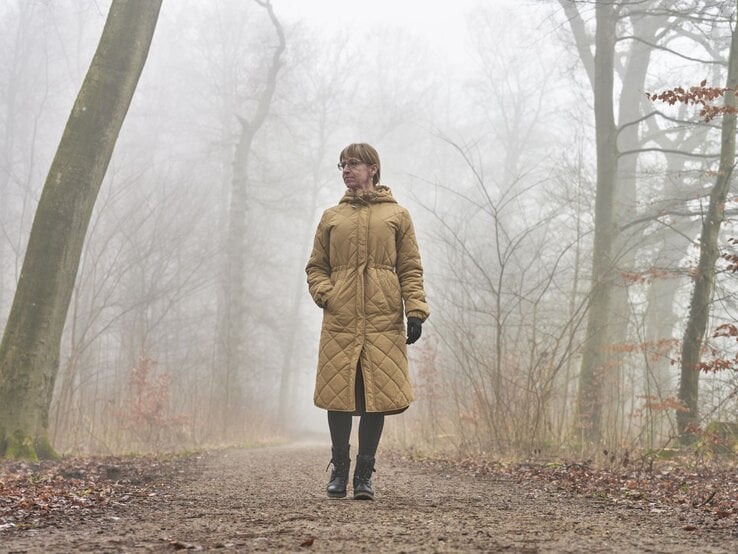 Ältere Frau mit braunem Steppmantel und Handschuhen spaziert nachdenklich auf einem nebligen Waldweg im Herbst.