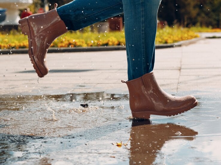 Nahaufnahme von Beinen in Jeans und glänzenden braunen Stiefeln, die schwungvoll durch eine Pfütze auf einem sonnigen Weg treten.