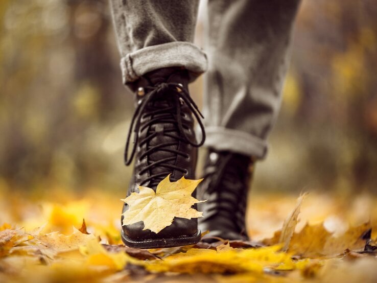 Ein gelbes Ahornblatt klebt an der Spitze eines schwarzen Stiefels, während eine Person durch buntes Herbstlaub geht.