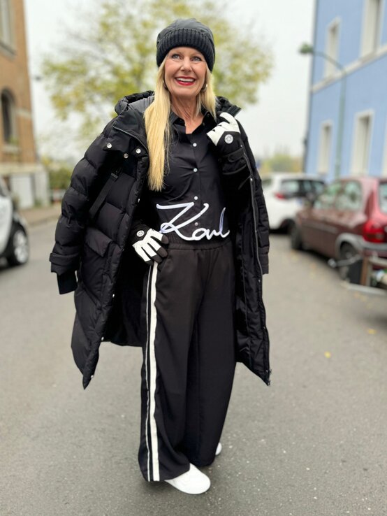 Eine blonde Frau in schwarzem Outfit mit gestreiften Hosen, dicker Steppjacke und Mütze lächelt auf einer Straße vor herbstlichem Baum. | © Renate Zott