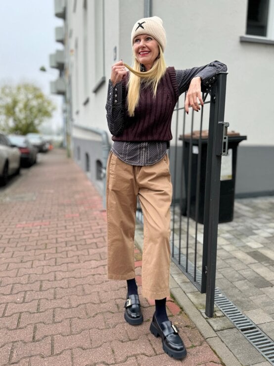 Eine blonde Frau in beiger Mütze, gepunkteter Bluse, braunem Pullunder und Stoffhose lehnt lächelnd an einem Metallzaun auf einem Gehweg. | © Renate Zott