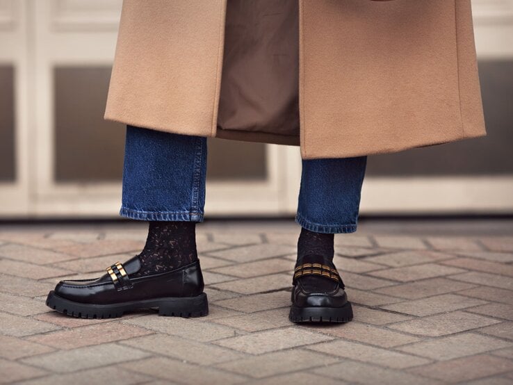 Schwarze Loafer mit goldenen Details und Spitzenstrümpfen, kombiniert mit hochgekrempelten Jeans und einem beigen Mantel auf Pflastersteinen.