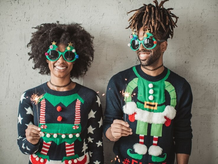 Zwei fröhliche Menschen in bunten Weihnachtspullovern und auffälligen Brillen mit Sternen und Weihnachtsdekor, halten Wunderkerzen vor grauer Wand.