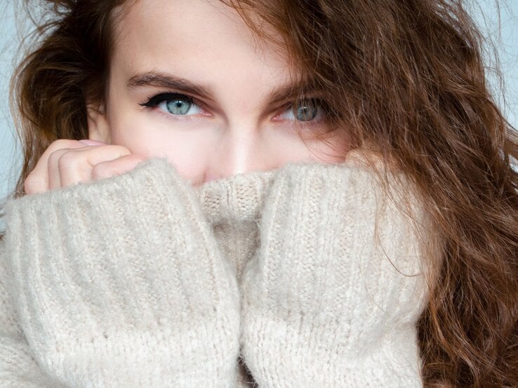 Junge Frau mit lockigem braunem Haar schaut mit hellblauen Augen über den Kragen ihres beigen Strickpullovers, nahes Porträt.