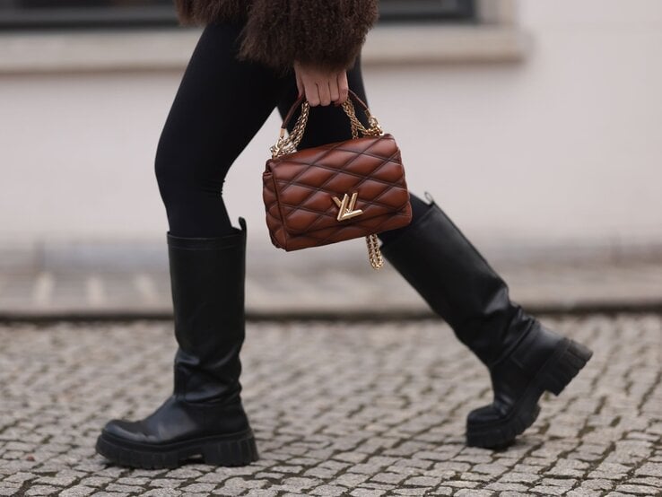 Nahaufnahme von Beinen in schwarzen Stiefeln auf Kopfsteinpflaster, eine braune Handtasche mit goldener Kette wird getragen.