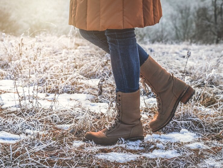 Detailaufnahme von Stiefeln und Jeans, die durch gefrorenes Gras und Schneeflecken in goldenem Morgenlicht schreiten.