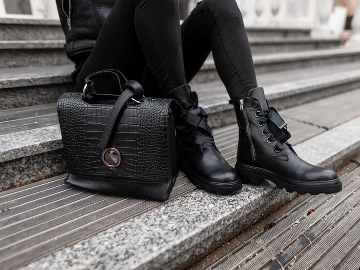 Nahaufnahme von schwarzen Lederstiefeln und einer luxuriösen Handtasche auf einer Treppe aus Stein und Holz.