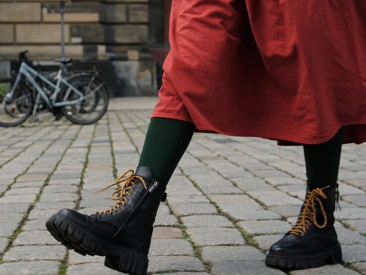 Schwarze klobige Dr Martens Stiefel mit senffarbenen Schnürsenkeln, getragen zu grün-schwarzen Strumpfhosen und einem roten Rock auf Kopfsteinpflaster.