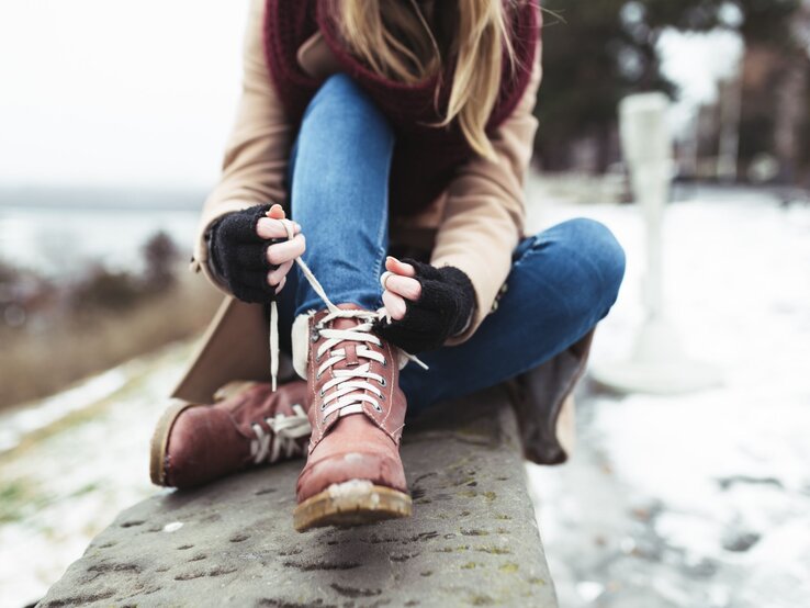 Eine Frau in Jeans und beigem Mantel schnürt braune Winterstiefel, sitzend auf einer Steinbank in verschneiter Landschaft.