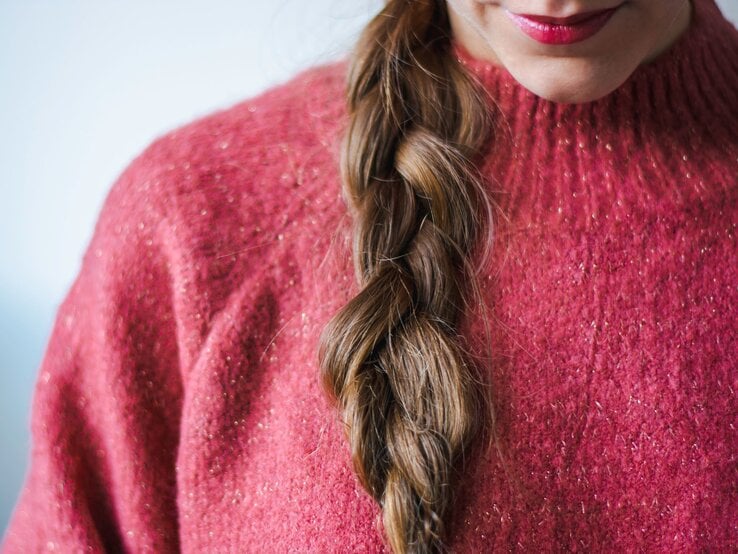 Eine Frau mit geflochtenem braunem Haar trägt einen rosafarbenen Glitzerpullover und dezenten Lippenstift.