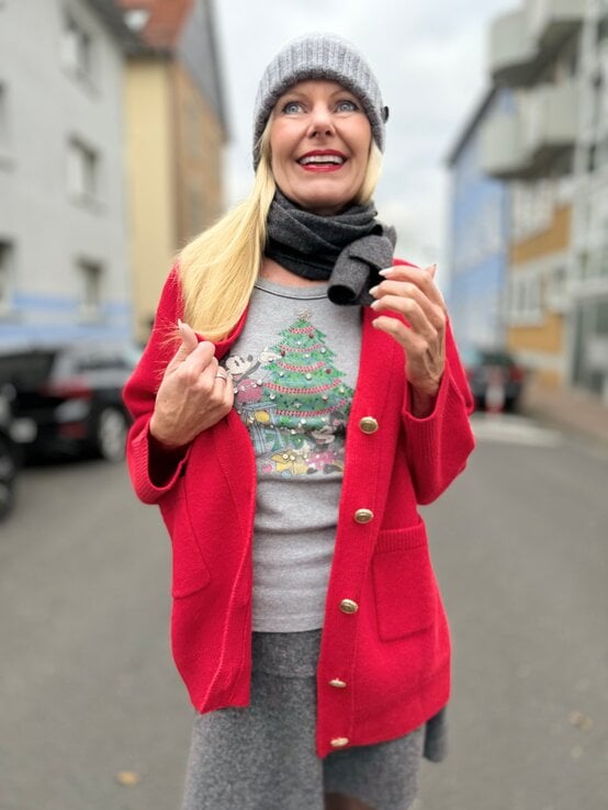 Eine blonde Frau mit grauer Mütze und Schal trägt eine rote Strickjacke und ein weihnachtliches Shirt, lächelnd auf einer Straße. | © Renate Zott