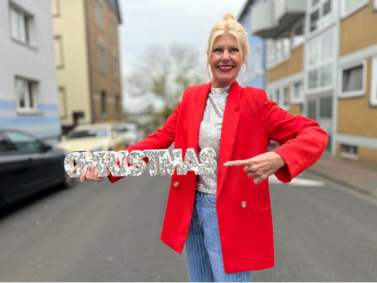 Eine blonde Frau in rotem Blazer und glitzerndem Shirt zeigt lächelnd auf eine silberne „Christmas“-Dekoration auf einer Straße. | © Renate Zott