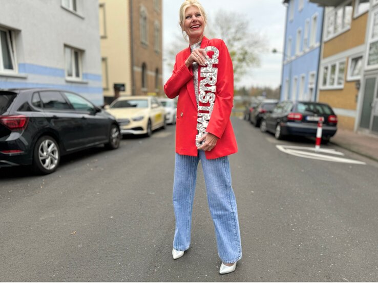 Eine blonde Frau in rotem Blazer und hellblauen Jeans lächelt auf einer Straße und hält eine silberne „Christmas“-Dekoration.