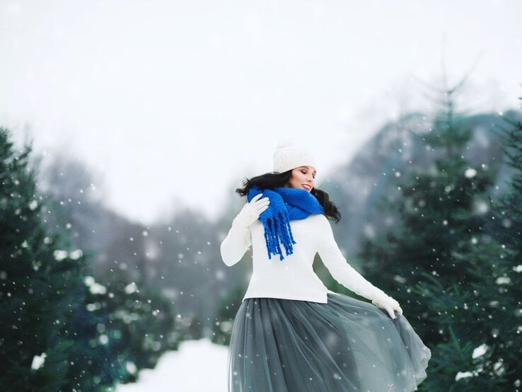 Frau mit hellem Wollpullover, grauem Tchibo-Tüllrock und blauem Schal dreht sich anmutig in einer schneebedeckten Waldlandschaft.