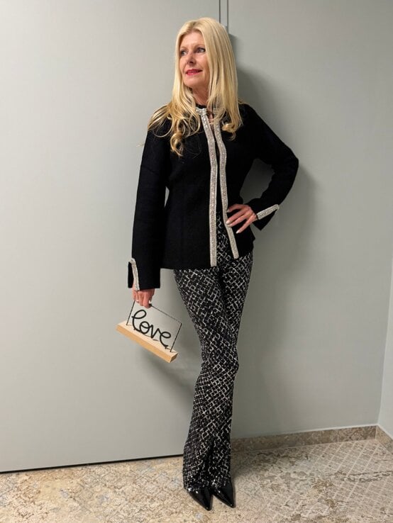 Eine blonde Frau in schwarzem, elegant verziertem Blazer und gemusterter Hose hält ein Schild mit der Aufschrift „Love“. | © Renate Zott