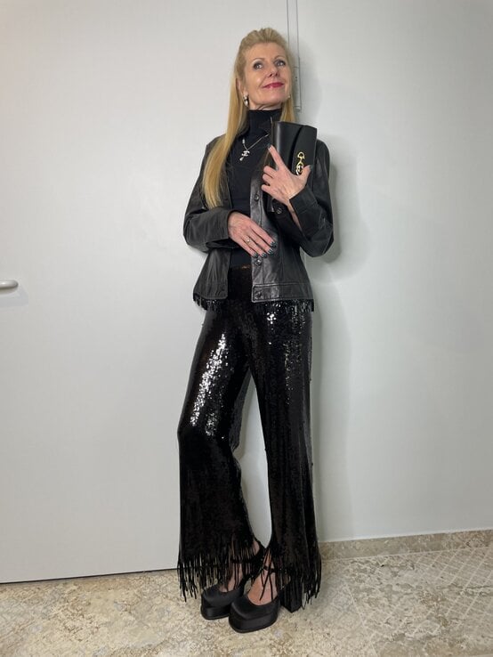 Eine blonde Frau in schwarzer Lederjacke, glitzernder Paillettenhose mit Fransen und schwarzen Plateau-Schuhen hält eine Clutch. | © Renate Zott