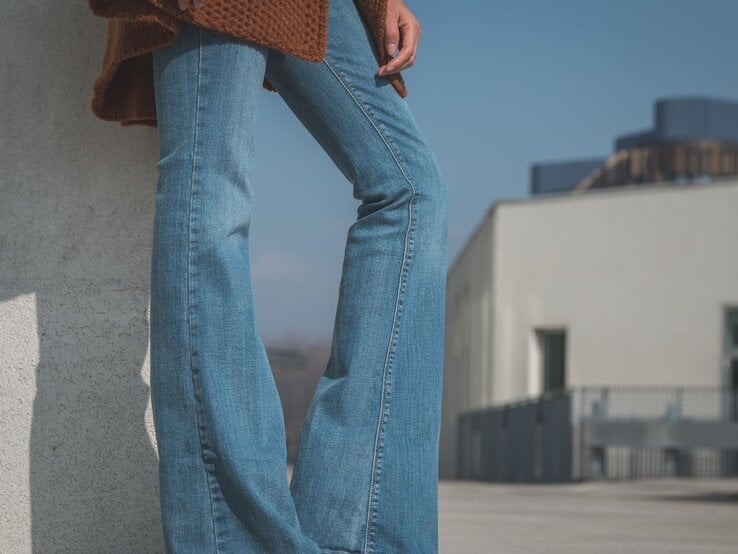 Detailaufnahme von blauer Schlaghose und braunem Strickoberteil, getragen vor urbanem Hintergrund bei klarem Himmel.