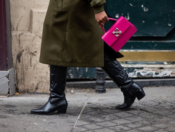 Schritt einer Frau in schwarzem Lederstiefel, olivgrünem Mantel und leuchtender pinker Tasche auf grauem Gehweg.