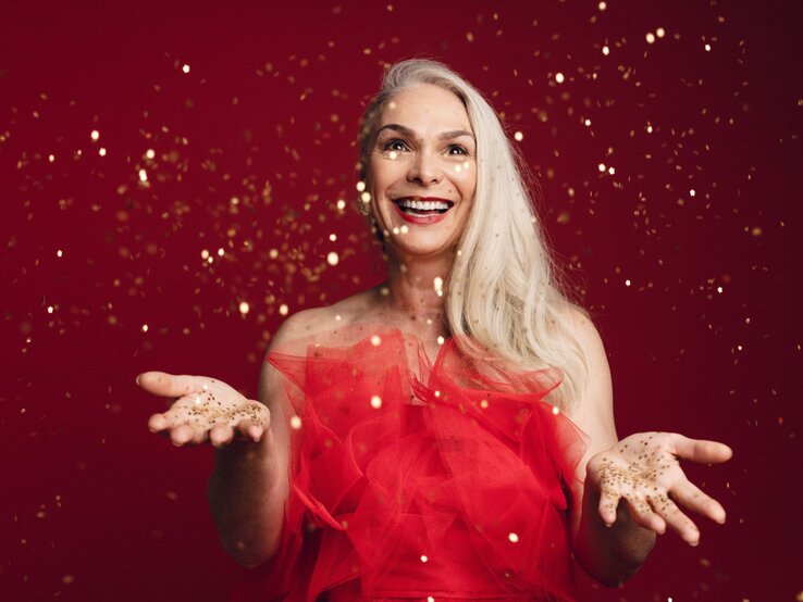 Strahlende Frau mit silbernem Haar in einem roten Kleid wirft goldene Partikel in die Luft, vor leuchtendem roten Hintergrund.