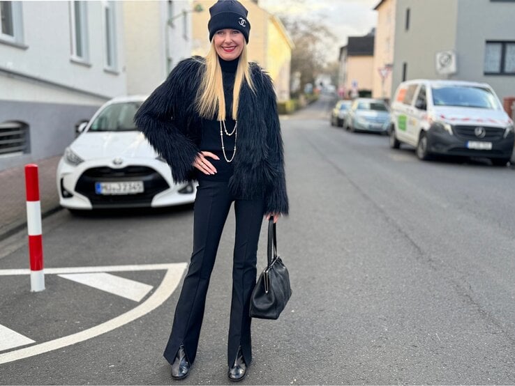 Eine blonde Frau in schwarzer Kleidung mit Pelzjacke, Mütze und Handtasche lächelt auf einer Straße mit geparkten Autos.