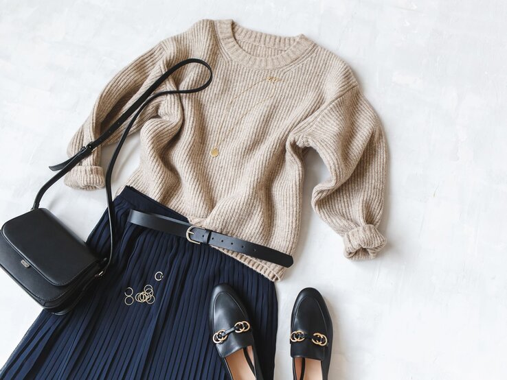 Elegantes Outfit aus beigem Pullover, dunkelblauem Strickrock und Accessoires wie goldenen Ringen, Tasche und Loafers auf weißer Fläche.