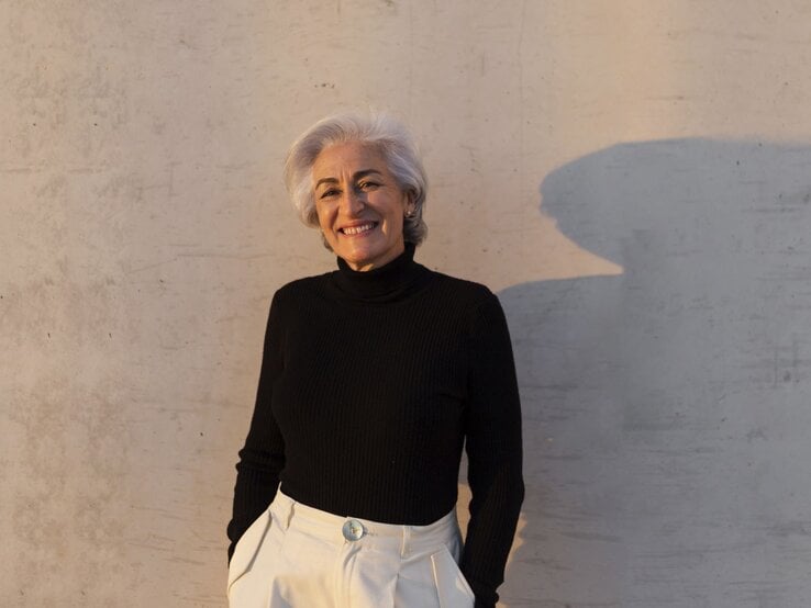 Elegante Frau mit grauem Haar in schwarzem Rollkragenpullover und heller Hose lehnt lächelnd vor einer Betonwand mit weichem Schatten.