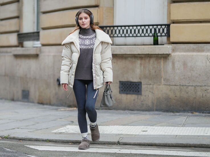 Frau mit beigem Wintermantel, grauem Pullover mit Schneeflockenmuster und Thermoleggings läuft konzentriert über einen Gehweg.