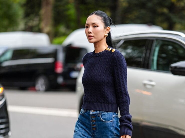 Elegante Frau mit glattem Zopf, dunkelblauem Pullover und hochgeschnittener Jeans geht konzentriert an parkenden Autos vorbei.