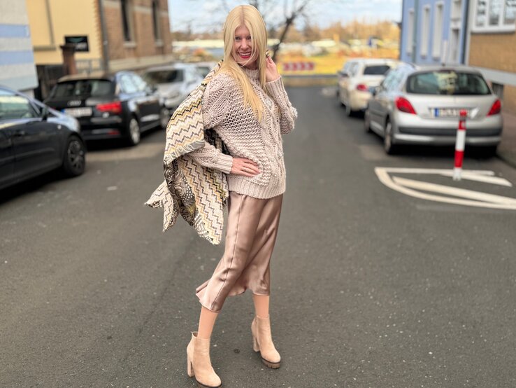 Renate Zott in beigem Strickpullover und glänzendem Satinrock posiert lächelnd in einer ruhigen Straße, umgeben von parkenden Autos. | © Renate Zott