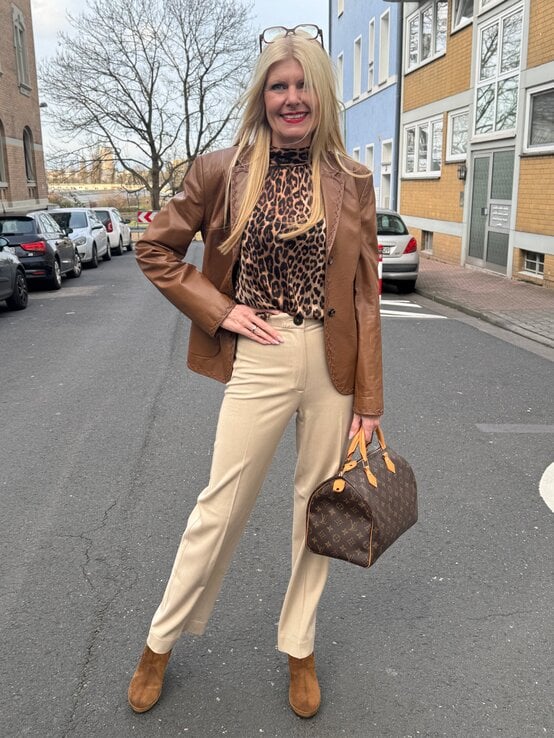 Renate Zott in brauner Lederjacke, Leopardenbluse und beigefarbener Hose hält eine Designer-Handtasche und posiert auf einer Straße. | © Renate Zott