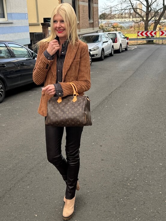 Reante Zott in braunem Cordblazer, schokobrauner Lederhose und beigefarbenen Stiefeletten trägt eine Designer-Handtasche auf einer Straße. | © Renate Zott