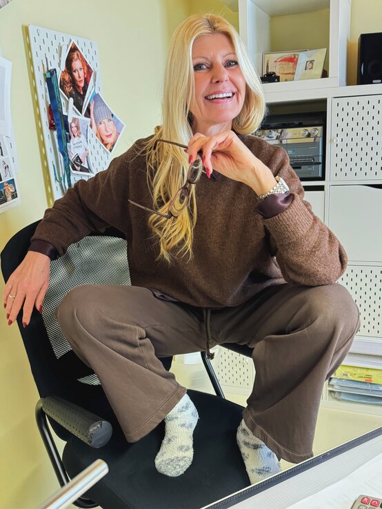 Freundlich lächelnde Renate Zott in braunem Outfit sitzt locker auf einem Bürostuhl, mit einer Pinnwand und Regalen im Hintergrund. | © Renate Zott