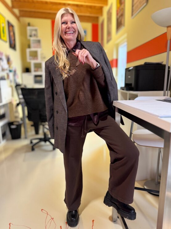 Lachende Renate Zott mit langen blonden Haaren, eleganter brauner Kleidung und klobigen schwarzen Schuhen posiert entspannt in einem modernen Büro. | © Renate Zott