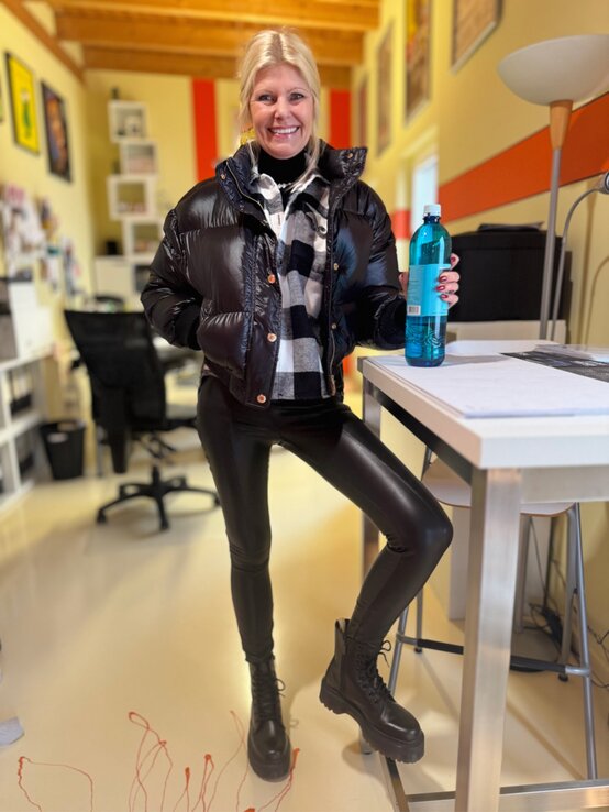 Blonde Renate Zott in schwarzer Daunenjacke, Karojacke und Lederleggings steht lächelnd an einem Tisch im Büro und hält eine blaue Wasserflasche. | © Renate Zott