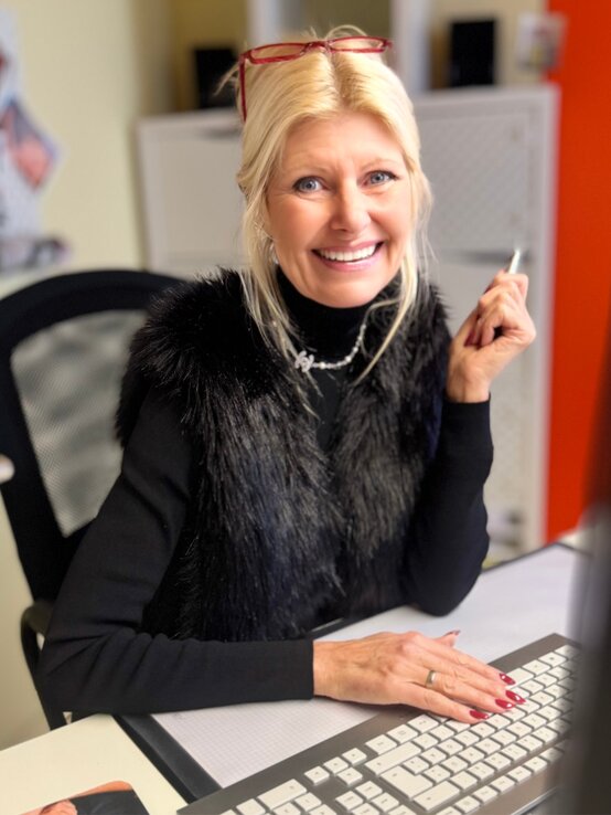 Lachende Renate Zott mit blondem Pferdeschwanz und elegantem schwarzen Look hält einen Stift in der Hand und arbeitet an einem Schreibtisch. | © Renate Zott