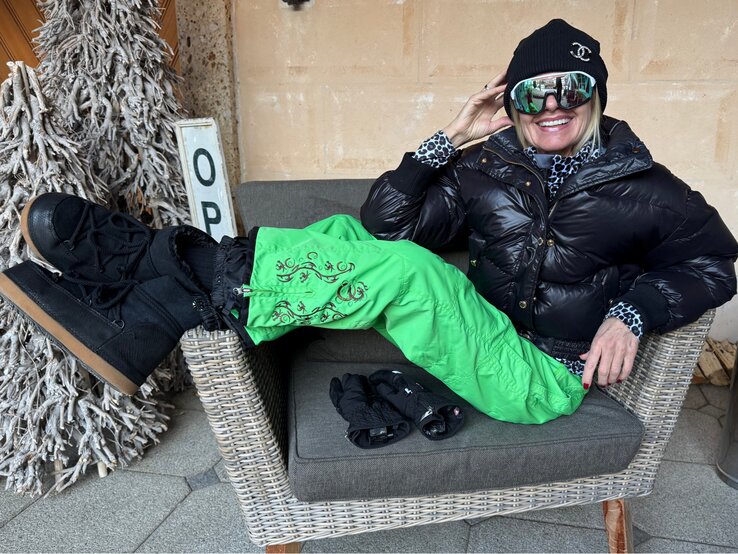 Renate Zott in schwarzer Daunenjacke, grüner Schneehose und dicker Mütze sitzt leger auf einem Korbstuhl, ein Handschuhpaar liegt daneben.