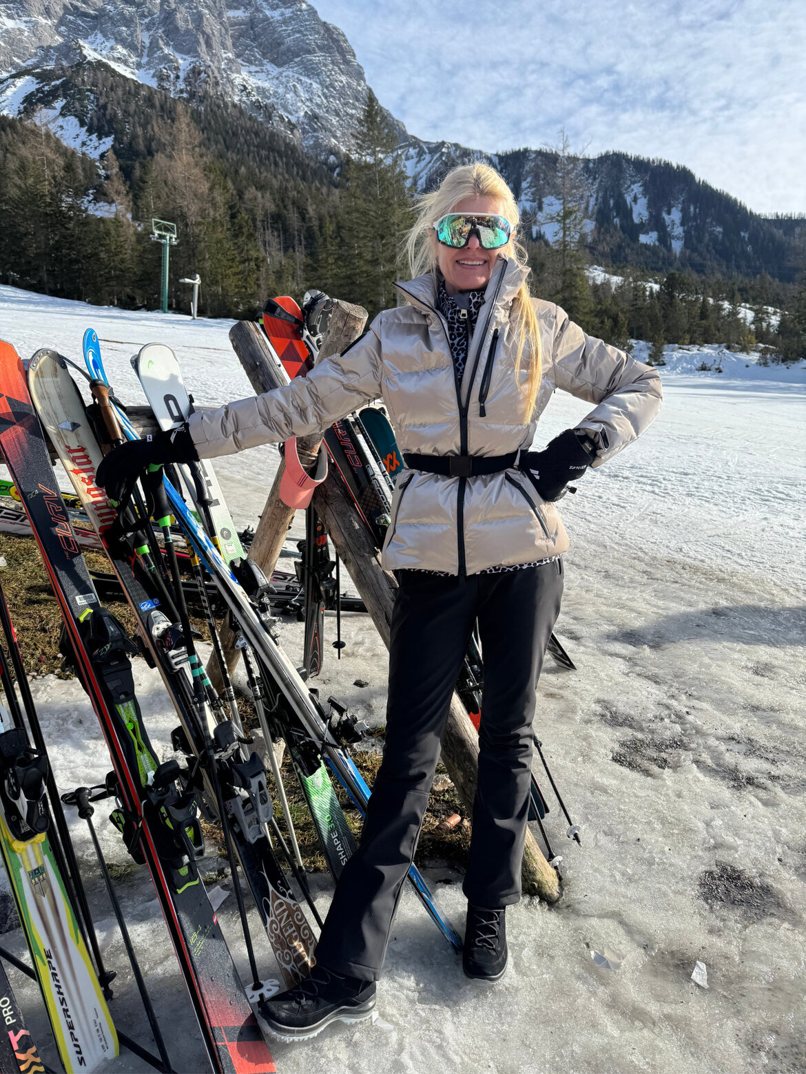 Renate Zott in silberner Daunenjacke, schwarzer Skihose und verspiegelter Skibrille lehnt lächelnd an einem Holzständer mit Skiern. | © Renate Zott