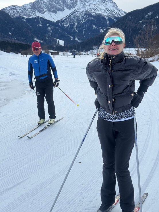 Renate Zott in schwarzer Daunenjacke und verspiegelter Skibrille steht lächelnd mit Langlaufskiern auf einer Schneeloipe vor Bergkulisse. | © Renate Zott