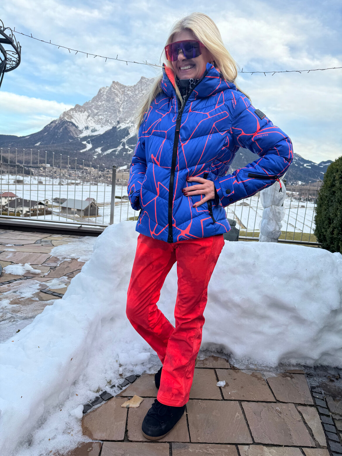 Renate Zott in blauer Daunenjacke mit roten Linien, roter Skihose und verspiegelter Brille posiert lächelnd vor verschneiter Bergkulisse. | © Renate Zott