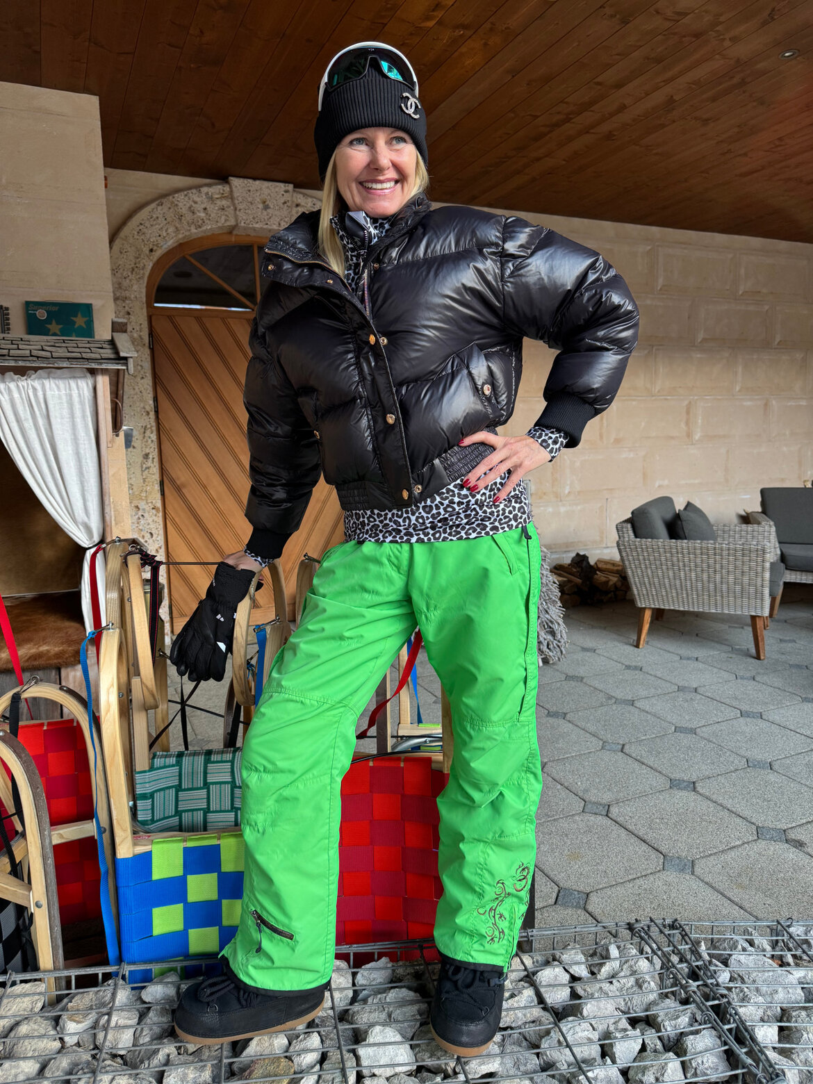 Renate Zott in schwarzer Daunenjacke, grüner Skihose und Mütze mit Logo posiert lachend vor Holzschlitten mit bunten Sitzpolstern. | © Renate Zott