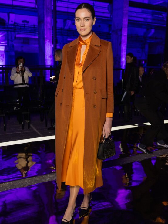 Elegante Gloria-Sophie Burkandt mit dunklem, zurückgebundenem Haar trägt ein orangefarbenes Satinkleid und braunen Mantel vor violett beleuchteter Kulisse. | © Andreas Rentz/Getty Images