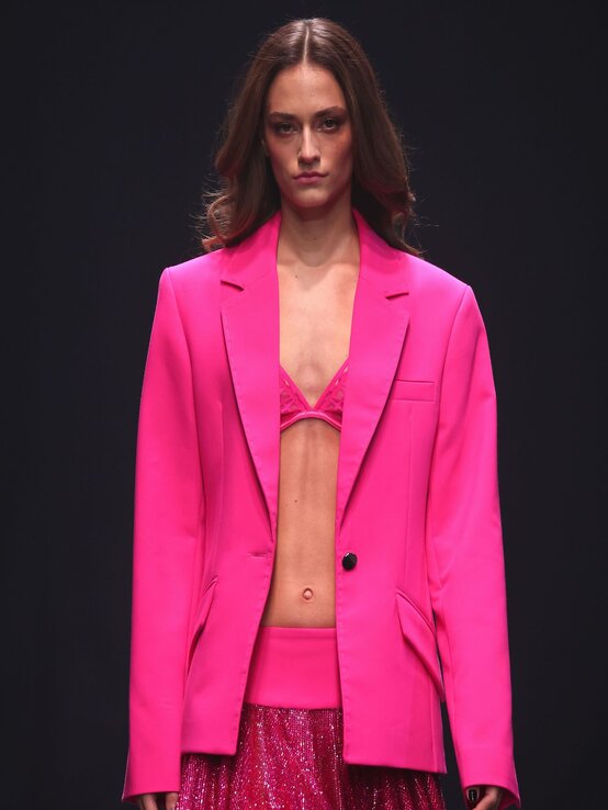 Model mit welligem Haar trägt einen oversized pinken Blazer über einem glitzernden Minirock und Spitzen-Bralette auf dem Laufsteg. | © Sebastian Reuter