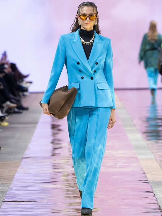 Eine Frau mit zurückgekämmtem Haar läuft in einem himmelblauen Anzug über den Laufsteg, trägt eine goldene Kette, gelbe Sonnenbrille und hält einen braunen Hut. | © Marc Cain 